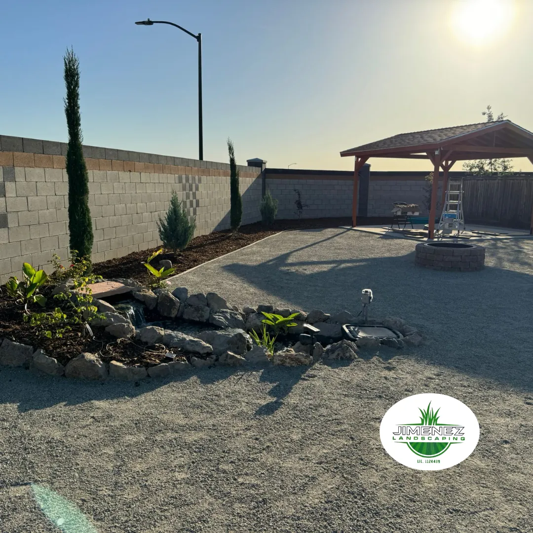 Landscape design featuring decorative rock, plants, and a stone water feature in a Bakersfield outdoor space, complemented by a wooden gazebo and a well-maintained gravel area. Jimenez Landscaping logo visible.