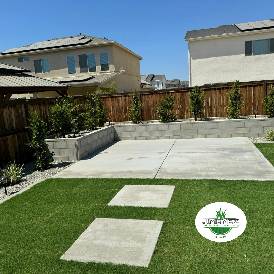 Concrete patio and walkways with green grass in a landscaped backyard, featuring decorative shrubs and a wooden fence, showcasing Jimenez Landscaping's concrete services in Bakersfield.