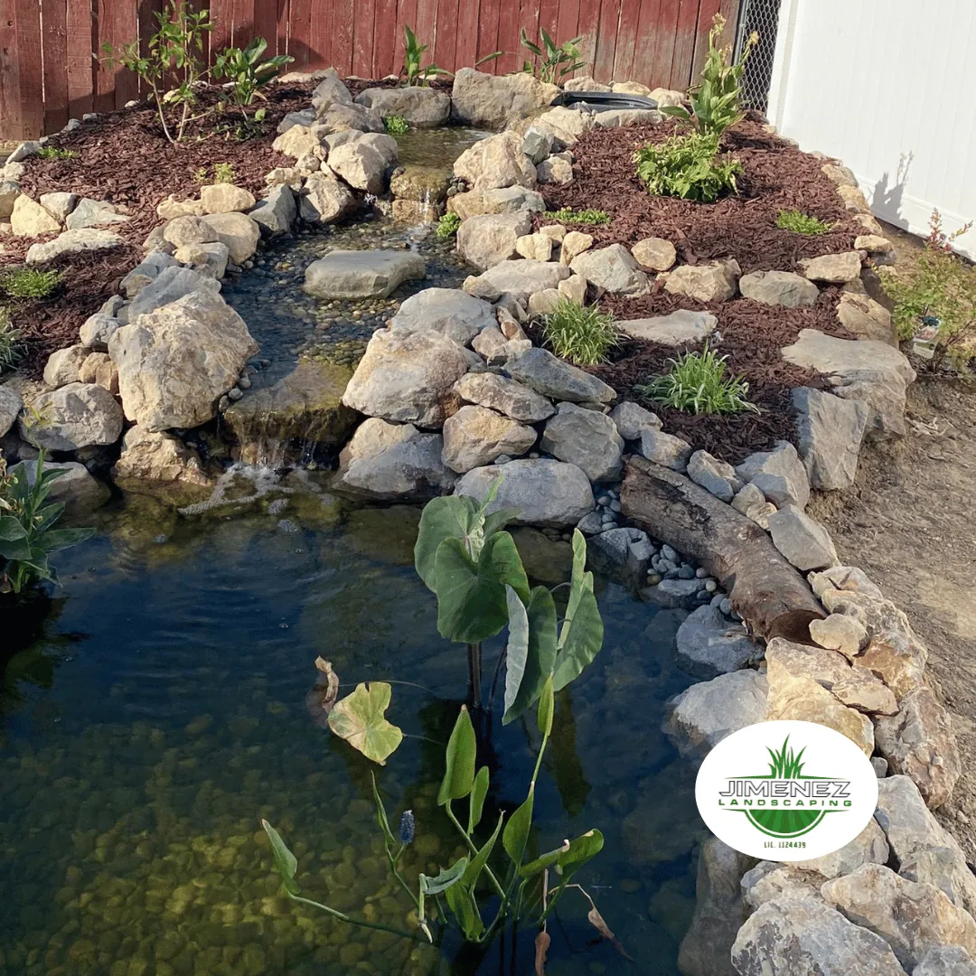 Rock-lined pond with a small waterfall and aquatic plants, featuring decorative mulch and landscaping elements, designed by Jimenez Landscaping in Bakersfield.