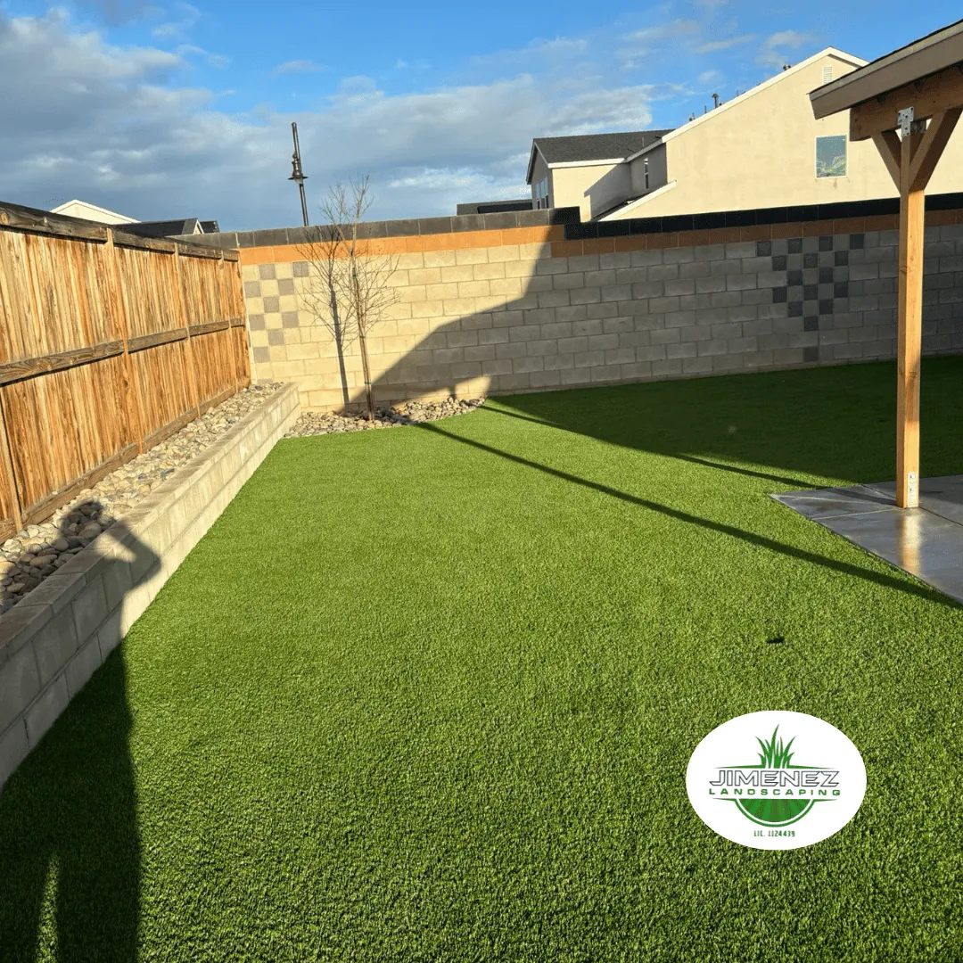 Artificial turf installation in a backyard with a wooden fence and decorative rocks, featuring Jimenez Landscaping logo, enhancing outdoor space in Bakersfield.